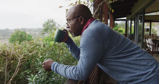 African american senior man standing on balcony drinking coffee and looking at view. retirement lifestyle, spending time alone at home.