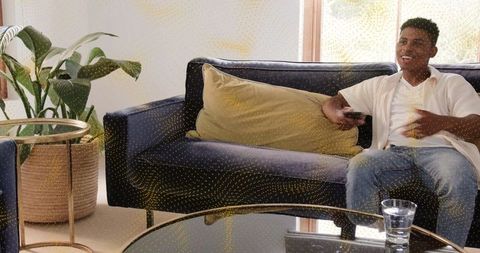 Young man relaxing on modern navy sofa holding remote smiling in bright cozy living room
