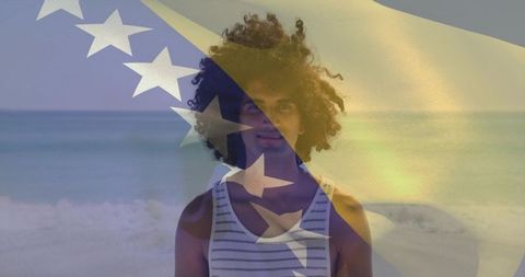 Curly-haired man standing on beach wearing striped tank with flag overlay at golden hour