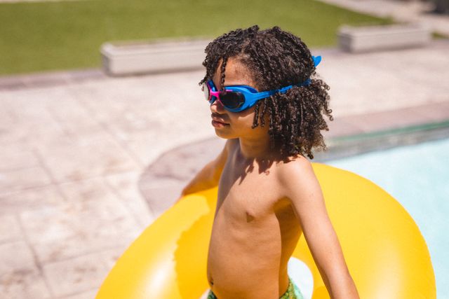 Shirtless hispanic boy wearing swimming goggles holding yellow inflatable tube at poolside. unaltered, childhood, enjoyment and summer weekend concept.