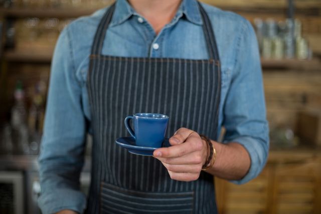 Barista in denim shirt and striped apron holding blue espresso cup on saucer. Ideal for concepts of coffee service, hospitality, barista training, cafes, morning routine, customer service, and food industry promotions.