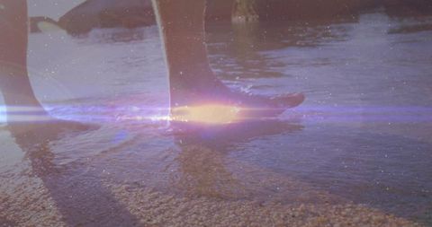 Sunlit barefoot man wading on pebbly shoreline with lens flare, calm shallow water