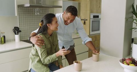 Couple sharing morning coffee at kitchen island while checking smartphone and laughing