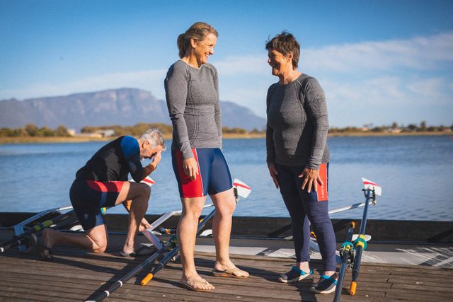 Two senior women and a man from a rowing club are preparing their equipment by the riverside. They are engaged in conversation and appear to be enjoying their active retirement lifestyle. This image can be used to promote healthy living, senior fitness, outdoor activities, and the benefits of staying active in later years.