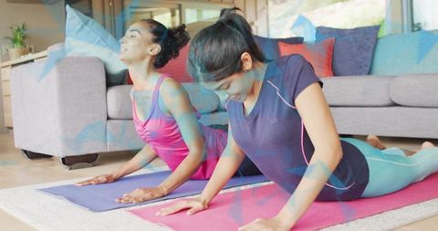 Two women practicing cobra pose on colorful yoga mats in sunlit living room