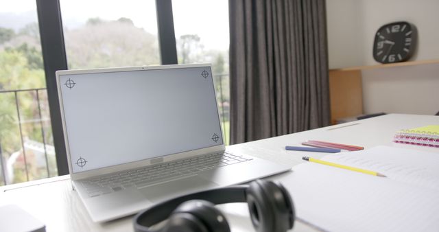 Laptop and black headphones placed on desk with various stationery items and notebook at home office. Beyond the desk is large window allowing natural sunlight into the room. Scene represents ideal workspace for remote work or study, promoting comfort and productivity.