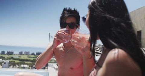 Couple sipping cocktails at rooftop pool edge with ocean view, sunglasses, summer terrace