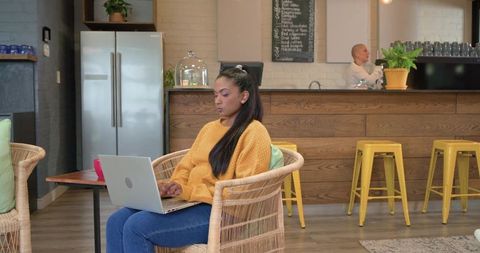 Young woman working on laptop in cozy cafe with rattan seating and yellow barstools