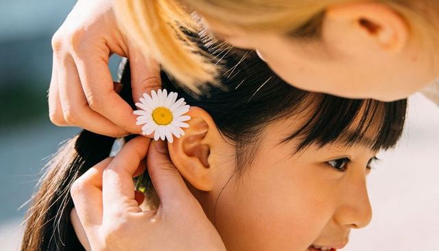 Placing daisy behind child's ear in sunlit park family bonding gentle outdoor moment