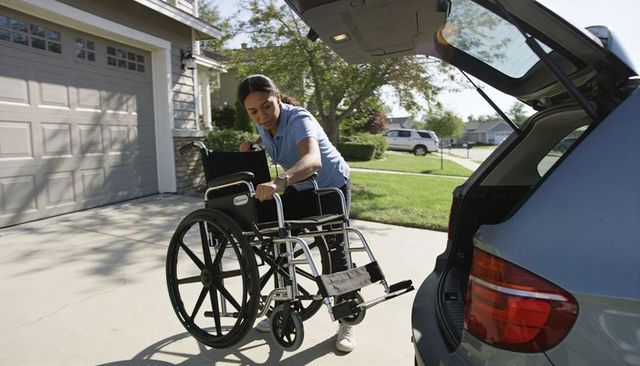 Caregiver loading manual wheelchair into suv hatch in suburban driveway for transport