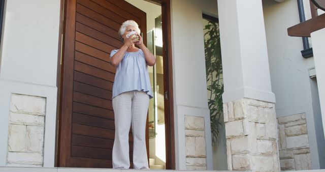 An elderly woman standing at the entrance of her home, holding a beverage, and taking in the fresh air. This image conveys a sense of relaxation, tranquility, and personal leisure time. Perfect for usage in articles or advertisements about senior wellness, lifestyle content focusing on serene home life, and promotional materials for retirement community benefits.