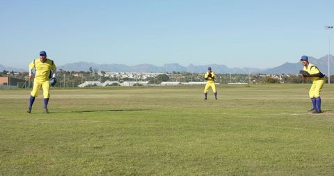 Three Outfielders Fielding on Sunlit Grass Outfield with Urban Skyline and Mountains