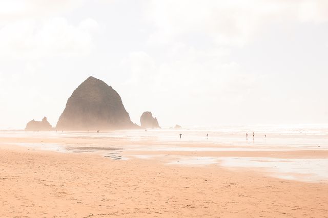 People walking along Cannon Beach in Oregon with Haystack Rock visible in the distance. The beach is sandy and the weather appears foggy, creating a serene and tranquil atmosphere. Perfect for travel blogs, vacation advertisements, nature and coastal scenery promotions, or to enhance tourism websites highlighting popular destinations.