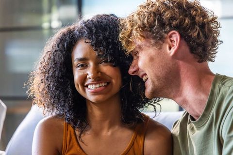 Laughing interracial couple sharing cozy couch moment, black woman smiling, white man leaning