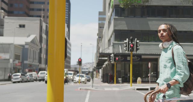 A young man with braided hair is crossing a city street on a sunny day. He is wearing headphones around his neck and carrying a backpack, suggesting he may be a student or commuter. The cityscape features tall buildings and traffic lights, capturing everyday urban life. Ideal for use in urban lifestyle, travel, or commuting-related content.