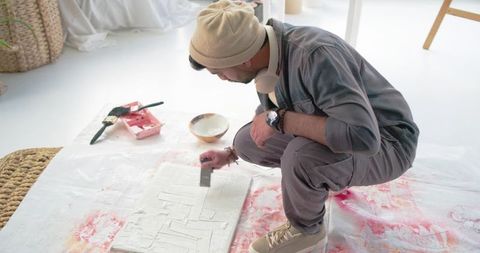 Indian artist scraping textured paste on canvas while crouching in bright daylight studio
