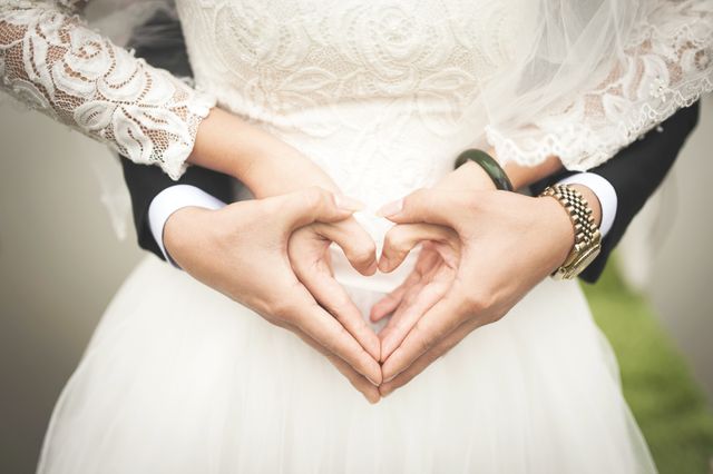 The bride and groom hold hands together in the shape of a heart, highlighting love and unity in a wedding setting. The bride wears a detailed lace dress, emphasizing elegance and commitment. This image can be used for wedding invitations, romantic greeting cards, wedding-themed advertisements, or articles about marriage and relationships.
