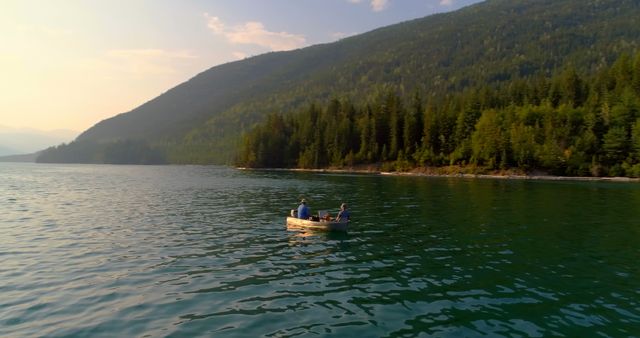 Two people are enjoying a peaceful boat ride on a serene lake surrounded by lush forests and mountains during sunset, with copy space. Their leisure activity amidst the natural beauty suggests a moment of relaxation and connection with nature.