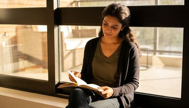 Sunlit indian woman reading on wide window ledge, cozy contemplative balcony moment