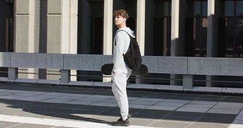 Young man standing on rooftop holding skateboard and looking upward in sunlit urban plaza