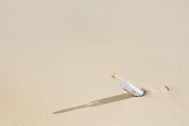 A lone bottle, partially buried in the sand, casting a shadow on a deserted beach. The bottle suggests themes of mystery, adventure, and isolation. Perfect for use in travel promotions, storytelling, adventure themes, or symbolic representations of communication and discovery.