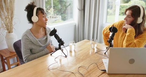 Multiracial women recording podcast at sunlit wooden table with microphones and laptop