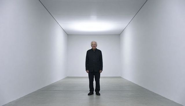 Solitary senior man standing in minimalist white gallery under soft ceiling light