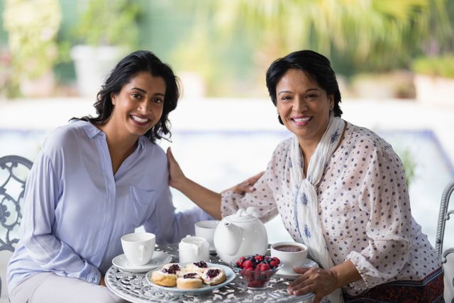 Mother and daughter sitting at a garden table, enjoying breakfast together. They are smiling and sharing a moment of togetherness with tea, pastries, and fresh berries. Ideal for use in family, lifestyle, and home-related content, emphasizing family bonds and morning routines.