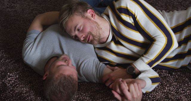 Happy caucasian gay male couple holding hand and looking to each other on carpet in living room. Togetherness, relationship, romance and domestic life, unaltered.