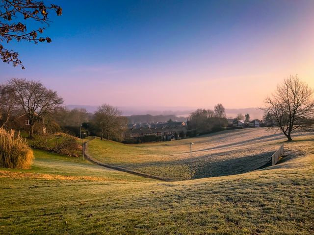 This image captures a frosty morning in a tranquil countryside with the first light of sunrise illuminating the serene winter landscape. Frost-covered grass, scattered trees, and a peaceful village with houses are visible in the background. There is a winding path leading through the grass and a wooden fence that complements the peaceful morning aura. This image is perfect for use in winter landscapes promotions, rural lifestyle illustrations, and calming morning scenes in prints and digital platforms.