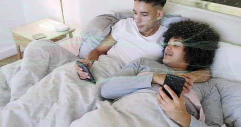 Male couple cuddling in bed using smartphones and tv remote under cozy neutral duvet