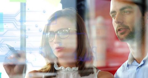 Woman pointing at circuit interface on glass with coworker using stylus in tech office