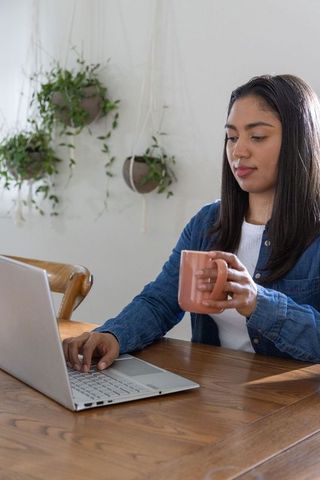Asian woman working from home with laptop and mug at sunlit wooden desk, hanging plants
