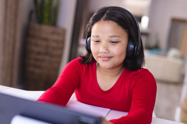 Asian girl wearing headphones sitting at desk using tablet, smiling. Ideal for educational content, technology use in learning, inclusivity in education, and lifestyle blogs. Perfect for illustrating remote learning, online classes, and children's use of technology.
