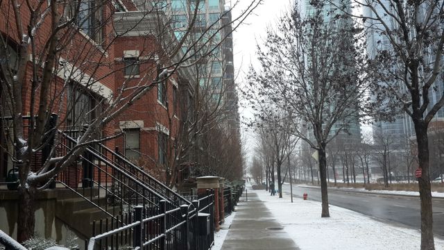 Winter street scene showcasing a snow-covered sidewalk bordered by leafless trees and red brick buildings on a cloudy day. Suitable for urban winter scenes, residential real estate promotions, seasonal backgrounds, and cityscape-related projects.