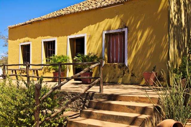 Charming yellow rural house with traditional architecture features rustic wooden fence and potted plants on a sunny day. Ideal for use in real estate promotions, tourism brochures, or articles about rustic living.