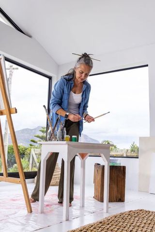 Senior woman artist standing in bright home studio arranging paintbrushes near wooden easel
