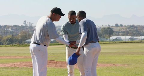 Diverse baseball team huddling on infield mound holding blue cap, teamwork and strategy