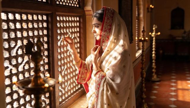 Indian woman gazing through carved jali window in cream saree with brass lamps