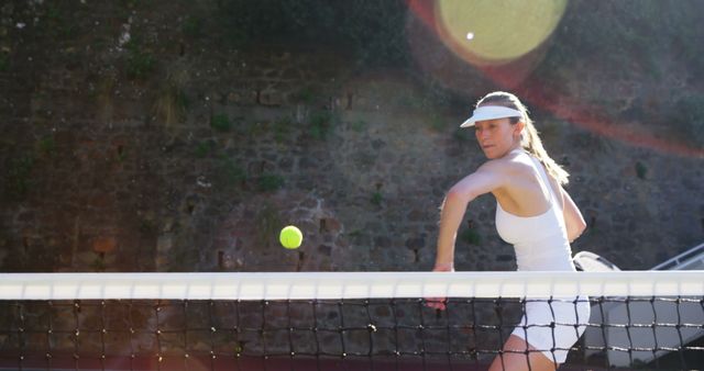 Woman playing tennis, returning ball with focused determination. Sunlight creates lens flare, adding drama to moment. Ideal for promoting sportswear, fitness, health campaigns, or competitive spirit. Great visual for sports events, posters, or tennis club promotions.