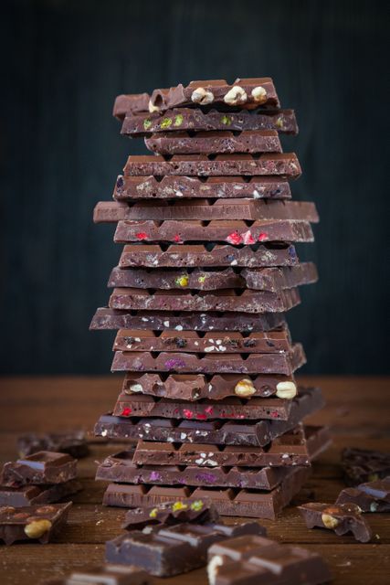 Tall stack of assorted chocolate bars with nuts and berries on wooden table, emphasizing variety and texture. Ideal for dessert blogs, confectionery advertisements, gourmet chocolate branding, and food photography portfolios.