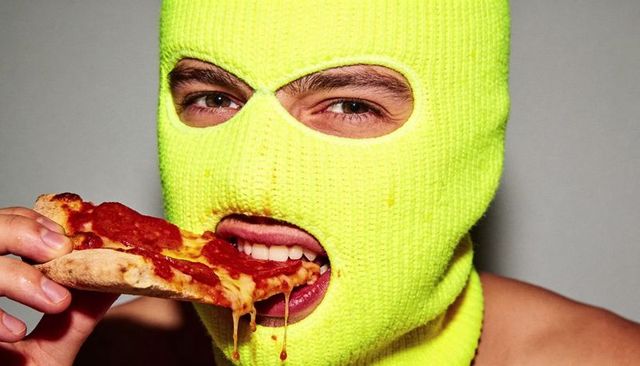 Shirtless man wearing neon yellow balaclava eating pepperoni pizza with melting cheese