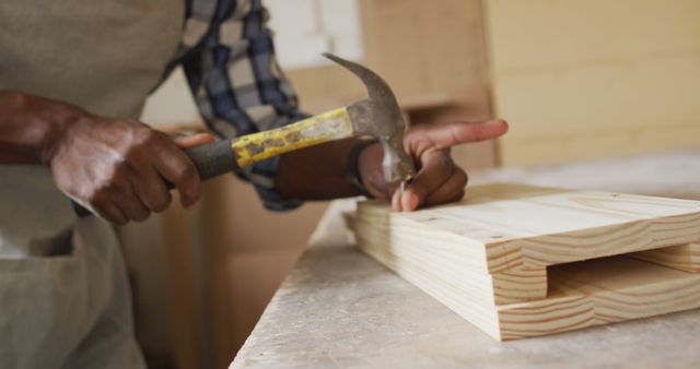 Mid section of african american male carpenter hammering nails into the wood at a carpentry shop. Carpentry, craftsmanship and handwork concept