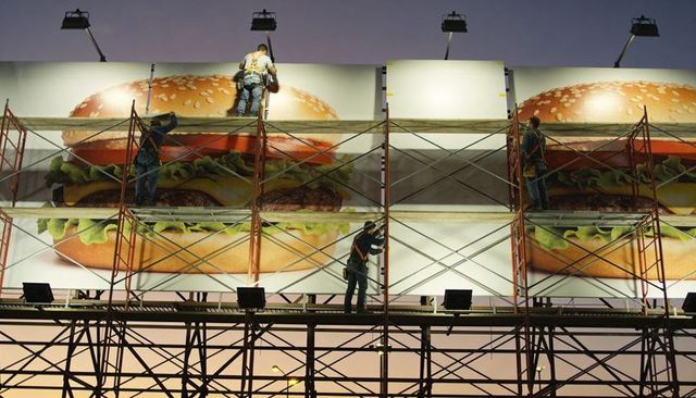 Construction crew installing giant burger billboard on scaffolding at dusk