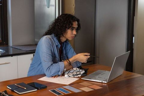 Non-binary designer reviewing fabric swatches on laptop while pointing pen at screen