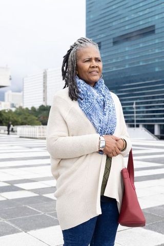 Senior woman checking smartwatch on plaza with blue highrise, patterned scarf and red tote