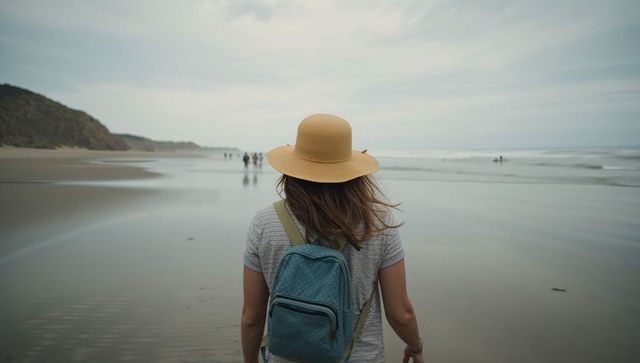 Woman walking along reflective beach at low tide wearing straw hat and teal backpack