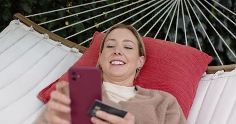 Mid-adult woman reclining on hammock using smartphone and credit card for online payment