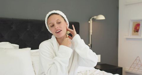 Woman massaging face with jade roller in white robe on bed morning skincare routine