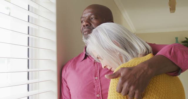 Senior diverse couple embracing and looking through window. Spending quality time at home and retirement concept.